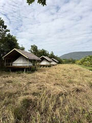 The image features a serene landscape with three elevated huts or cabins, each with thatched roofs and open sides, situated amidst lush greenery. 
