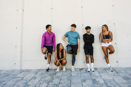 A female athlete sits next to a group of four athletes who are standing while they lean on a wall and talk