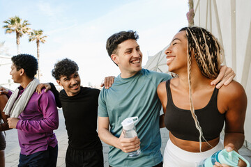 Male and female athletes are hugging and holding bottles while a male athlete walking next to them puts his hand on his shoulder and they laugh