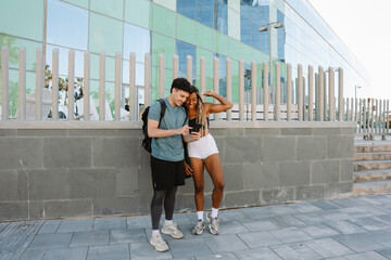Male athlete points at phone showing to female athlete as they stand and laugh while leaning on fence