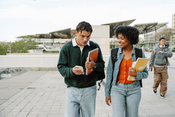 Female student smiling and listening to male student talking and showing phone while they walk and hold notebooks