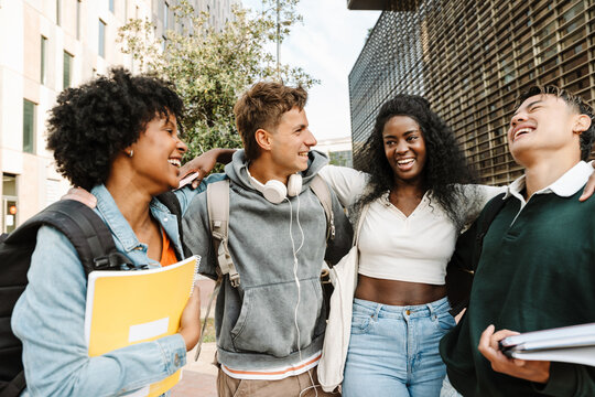 A group of four students stand and hug while laughing and two of them hold notebooks - Powered by Adobe