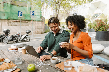 Female student holding a cup and sitting at a table with a male student while they laugh and look at a laptop