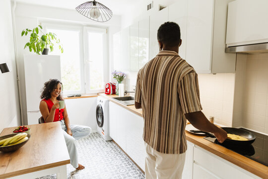 A woman sits at a table and holds a cup while smiling and talking to a man who is frying in a pan