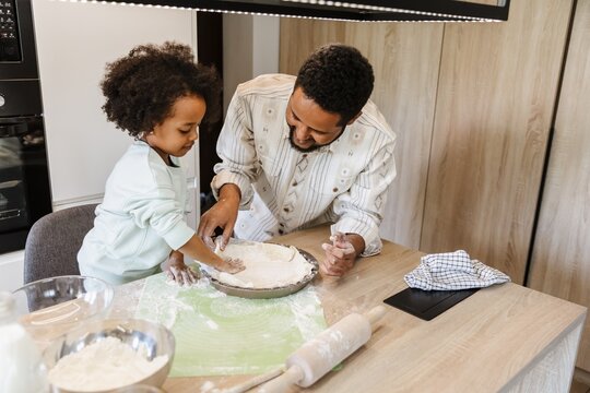 A father and his daughter are spreading dough on a baking sheet while standing at a table