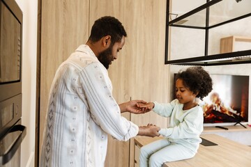 A daughter sits at a table and holds the hands of her father who is standing opposite her