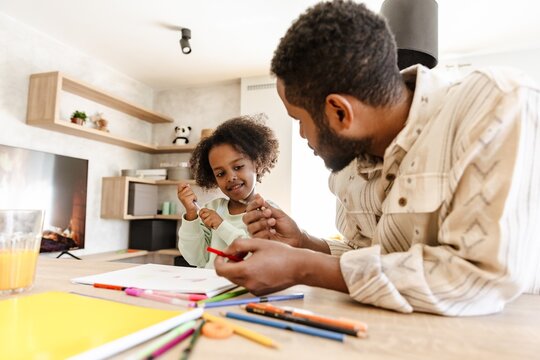 Daughter sits at the table and holds a pen while smiling and listening to her father talking and holding pencils