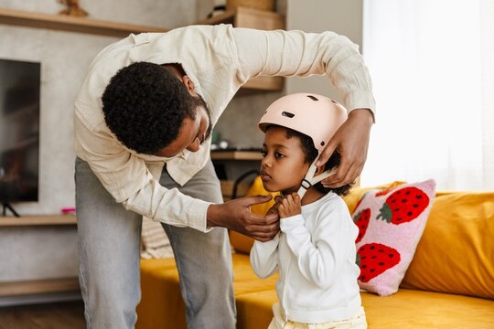Father stands leaning over and holding hands next to his daughter who is fastening her helmet