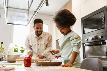 Daughter stands at the table holding a spoonful of sauce over a baking dish as her father...