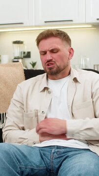 Young man at home presses hands to stomach feels cramps and pain after heavy fatty food and breathes. Caucasian guy on sofa curls slightly and shows nausea spasm and low mood while waiting for relief