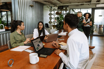 A female employee is talking to two coworkers while they are sitting at a table in front of laptops and two other coworkers are standing next to them
