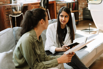 A female worker holds a notebook and pencil while listening to a female worker sitting next to her on a couch and holding documents