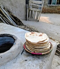 &ldquo;Fresh stack of homemade flatbreads placed on a traditional woven basket beside a clay tandoor oven in a rural village setting, mud house background, rustic outdoor kitchen, natural daylight, dry soil