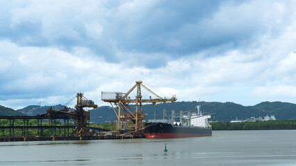 Bulk carrier ship moored at a rusty industrial loading terminal under a cloudy sky, surrounded by tropical hills and water