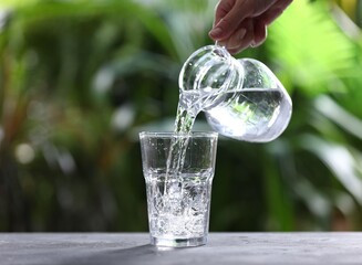 Woman pouring water into glass from jug at grey table against blurred green background, closeup