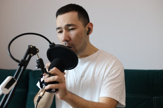 Asian man recording audio at home, speaking passionately into a microphone with raised hands. Podcasting, streaming, or voice-over session in a casual home studio