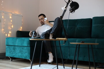 Asian man working from home on the sofa, multitasking with a phone call, tablet, and laptop. Casual remote-work setup with microphone, gadgets, and relaxed living-room environment
