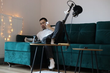 Asian man working from home on the sofa, multitasking with a phone call, tablet, and laptop. Casual remote-work setup with microphone, gadgets, and relaxed living-room environment