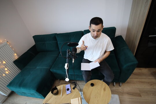 A man sits on a sofa in a home recording setup, holding notes and gesturing as he speaks into a microphone, with warm Christmas lights visible nearby