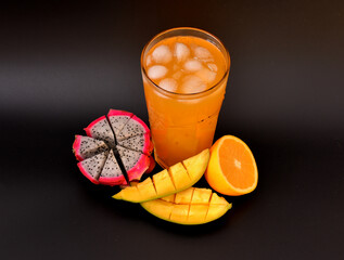 A tall glass of fruit juice with ice against a black background, next to pieces of ripe mango, dragon fruit, and orange.