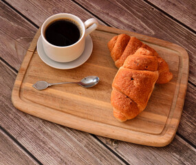 Wooden tray with a cup of black coffee on a saucer and two fresh croissants on a wooden table.
