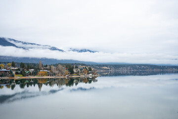 Calm lake reflects the houses and mountains under the cloudy skies above.