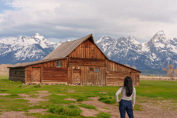 A woman traveler stands in front of a perfect blend of history and wilderness, with the Moulton Barn and stormy Teton Mountains.