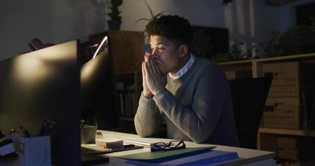 Working office worker wearing grey sweater at night office desk, with two monitors, lamp, notebooks