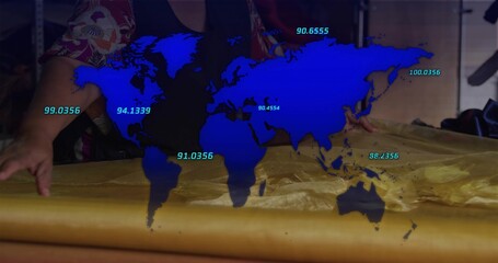 Leaning mature woman smoothing golden fabric on worktable, wearing floral shirt, blue map overlay