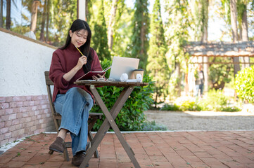 Asian woman holding pencil looking at book aside laptop while sitting at wooden table in garden cafe