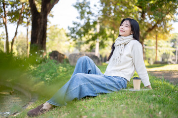 Asian woman in scarf leaning back sitting leisurely aside coffee on grass in garden or nature park.