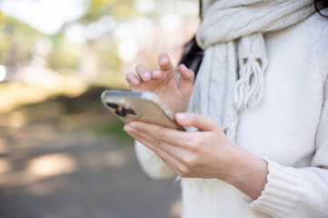Close up of woman in scarf holding and looking at phone as standing walking in garden or nature park