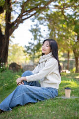 Obraz premium Asian woman in scarf sitting holding knee up aside coffee on grass field in garden or nature park.