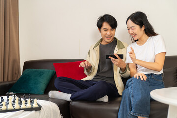 Young man lifestyle playing chess and using smartphone with woman sitting on sofa smiling