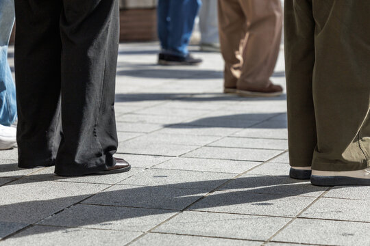 Diverse group of people standing on sunlit pavement cast shadows across tiles surface. Their jeans, khaki slacks, sneakers and casual shoes each tell a story of personal choice and expression.