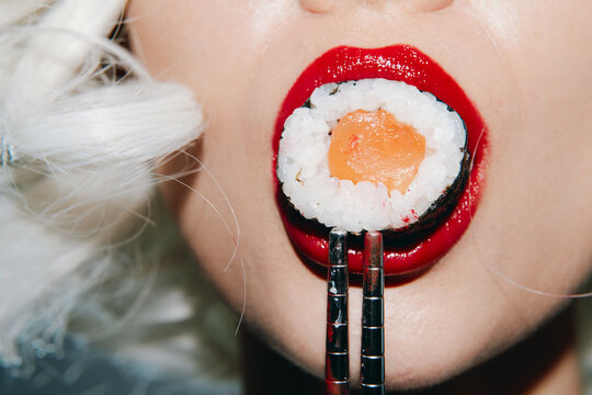 Close-up of a woman's mouth with red lipstick holding sushi, showcasing modern cuisine with a vibrant, playful aesthetic and bold colors.
