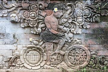 Stone sculpture on a temple wall in Singajara, Bali, Indonesia, depicting a man riding a bicycle. Created around 1906. The artwork notes the Dutch influence on the local culture. 
