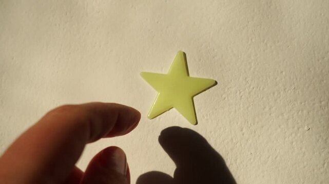 A hand gluing a luminescent five-pointed star, a self-adhesive toy, to a white wall. Decoration and interior design. Sunlight required to accumulate the luminescence effect