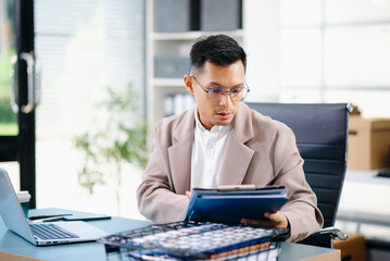 Obraz premium Confident Asian businessman standing at desk, reviewing report and working on laptop in modern office