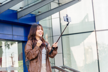 Smiling Asian woman, people enjoy shopping lifestyle fashion, posing with phone selfie stick for social media outside modern city mall building, capturing travel memory