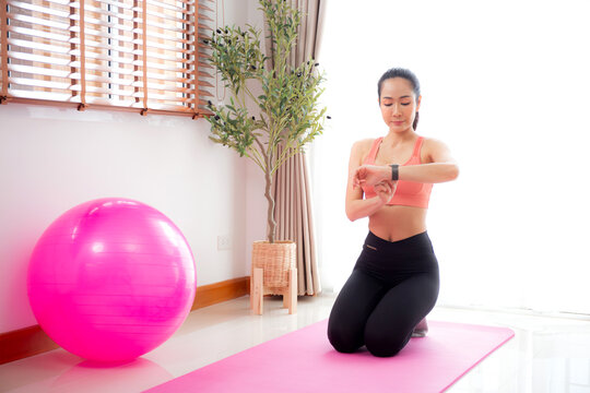 Young asian woman in activewear preparation practicing yoga while looking smartwatch sitting on yoga mat in living room at home, woman preparing workout with challenge and motivation, sport concept.