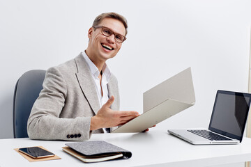 Smiling professional man with glasses wearing gray blazer working on laptop in modern office setting with smartphone and notebooks on desk, joyful business environment. People lifestyle concept.