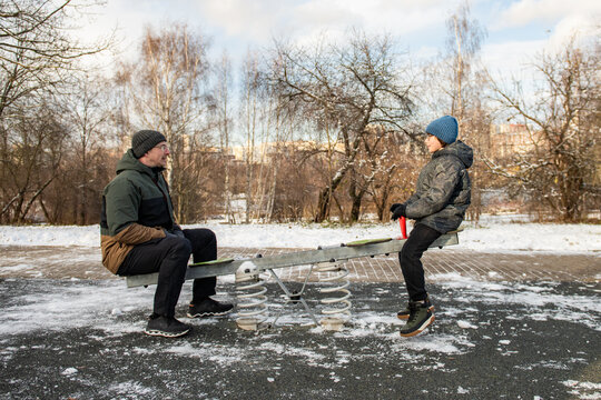 Adult and child sit on a seesaw in a snowy urban park, wearing winter coats and hats.