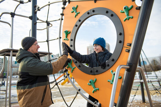 On a winter day, father and son high-five each other through a round hole in a colorful climbing structure on an outdoor playground. - Powered by Adobe