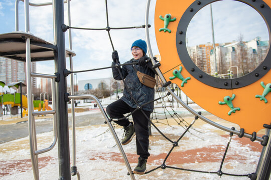 In winter, a boy climbs a rope net and a round game board to the city playground wearing a blue cap, a soft jacket, gloves, black trousers, green shoes and a small brown shoulder bag.