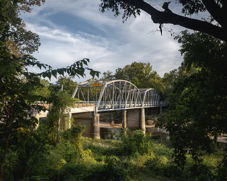 A scenic view of the 1923 Devil's Elbow Bridge spanning the Big Piney River in the Missouri Ozarks. The historic steel truss bridge is framed by lush green trees and foliage under a cloudy blue sky.