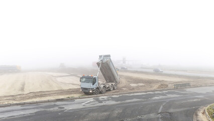 Dump truck unloading material on foggy construction site, showing industrial process, reduced visibility, working environment, and real heavy-duty operation in muted atmosphere.