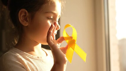 Young Girl Holding Gold Ribbon near Window in Bright Sunlight to Promote Childhood Cancer Awareness on a Sunny Day in Childhood Home Portrait - Powered by Adobe