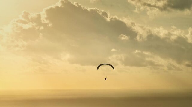 Paraglider soaring above calm ocean at golden-hour sunset over Bali cliffs, dramatic clouds and warm light conveying freedom, adventure and serene aerial landscape vistas