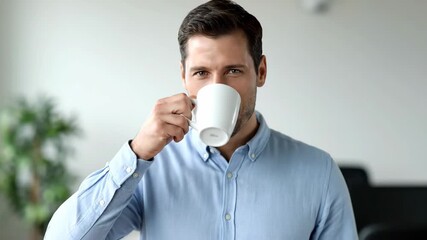 Man Enjoying a Coffee Break - A handsome man in a light blue shirt takes a sip from a white mug, looking directly at the camera with a slight smile.